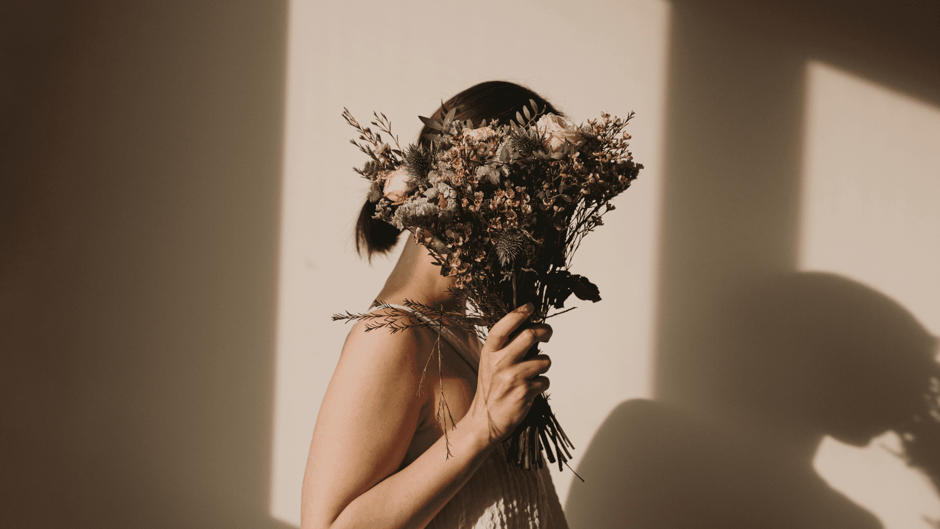 A person holding a bouquet, obscuring their face against a sunlit wall.